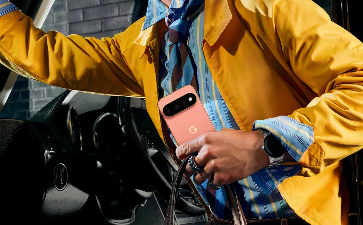 A person in a yellow jacket holds a coral Pixel 11 Pro phone while getting out of a car