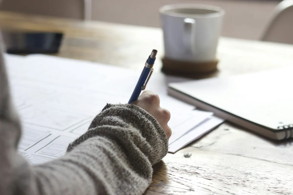 A person writes notes at a desk beside a laptop and coffee mug