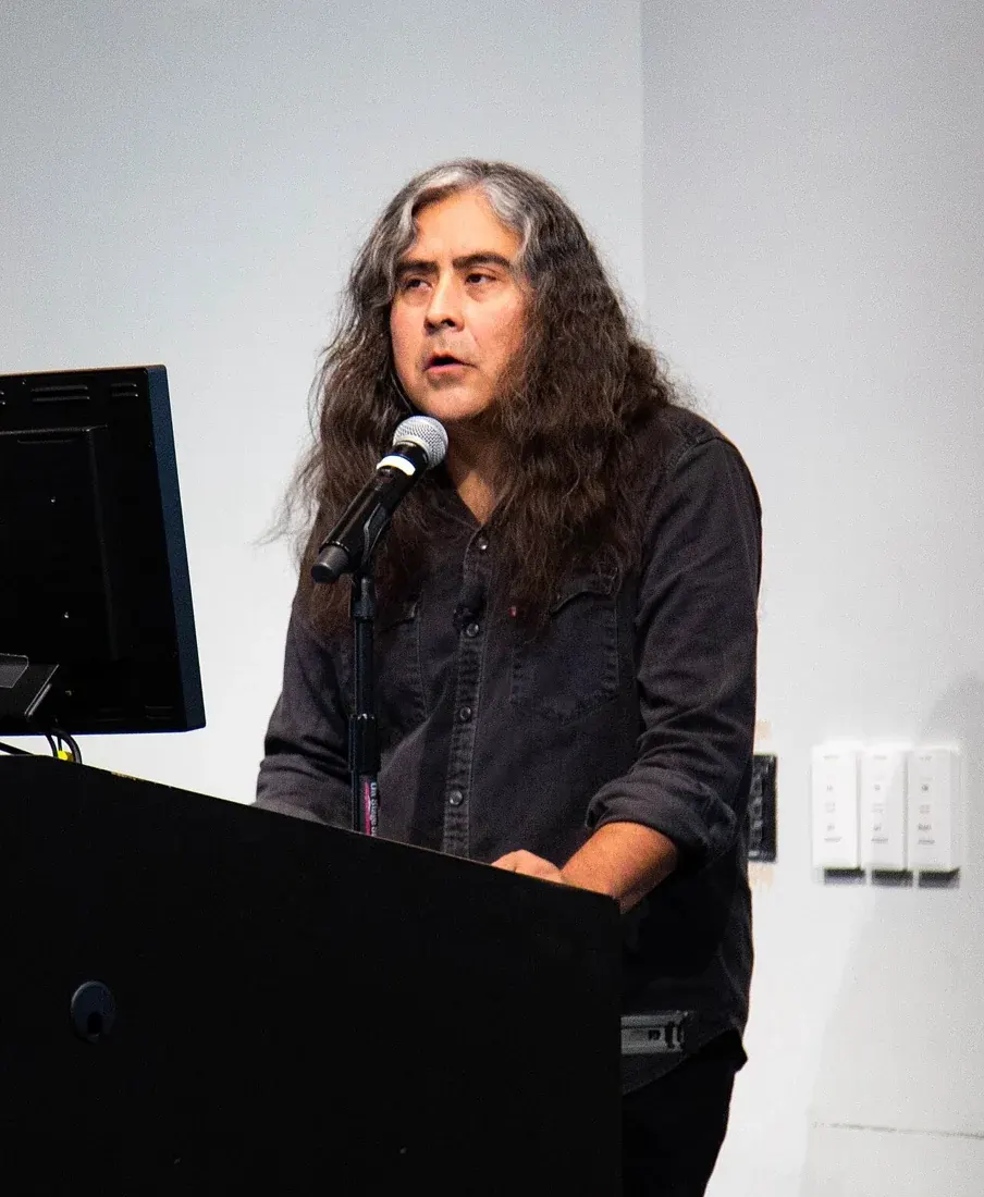 Raven Chacon speaks into a microphone in front of an audience
