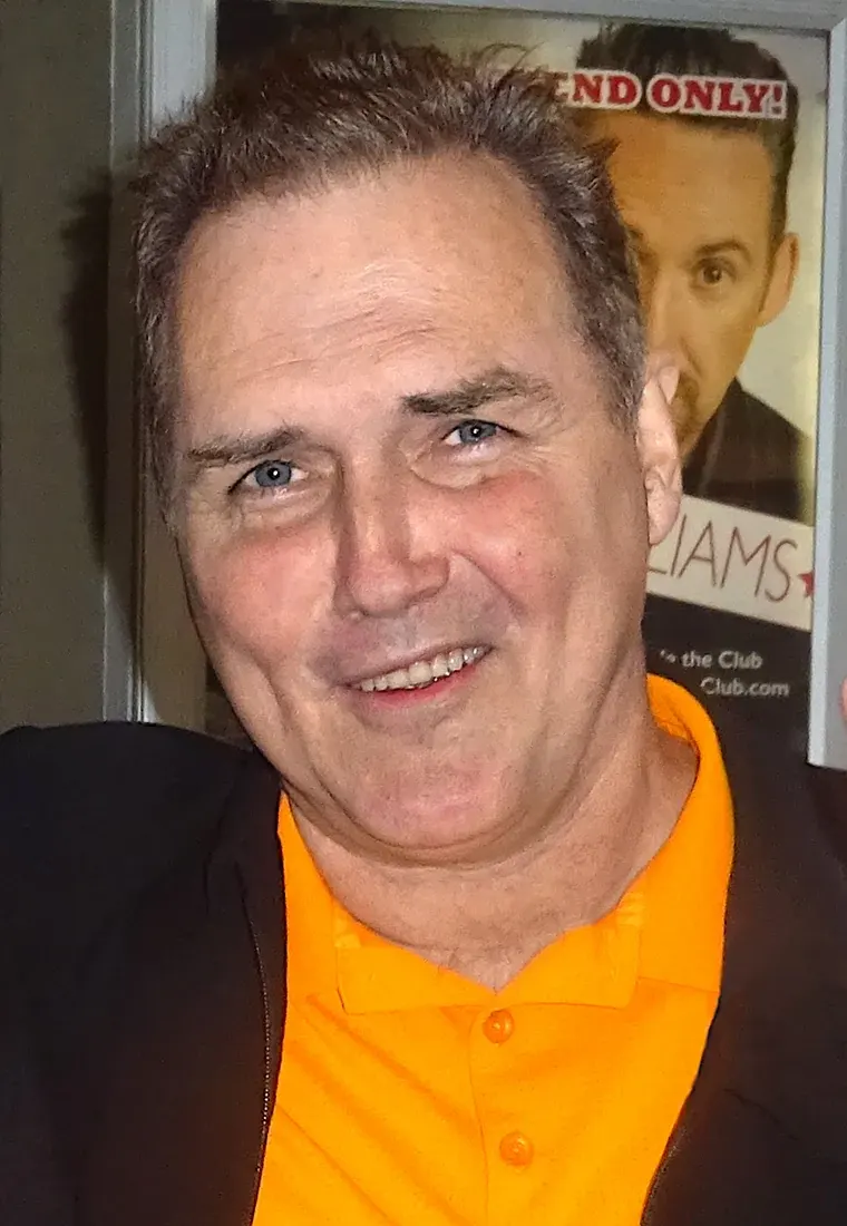 Norm Macdonald smiling in a yellow shirt and dark jacket