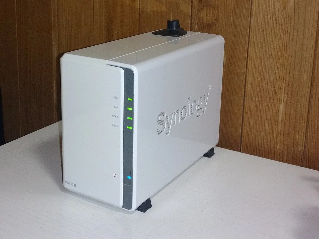 White Synology DS213j NAS with front status lights on a white surface against a wood wall.