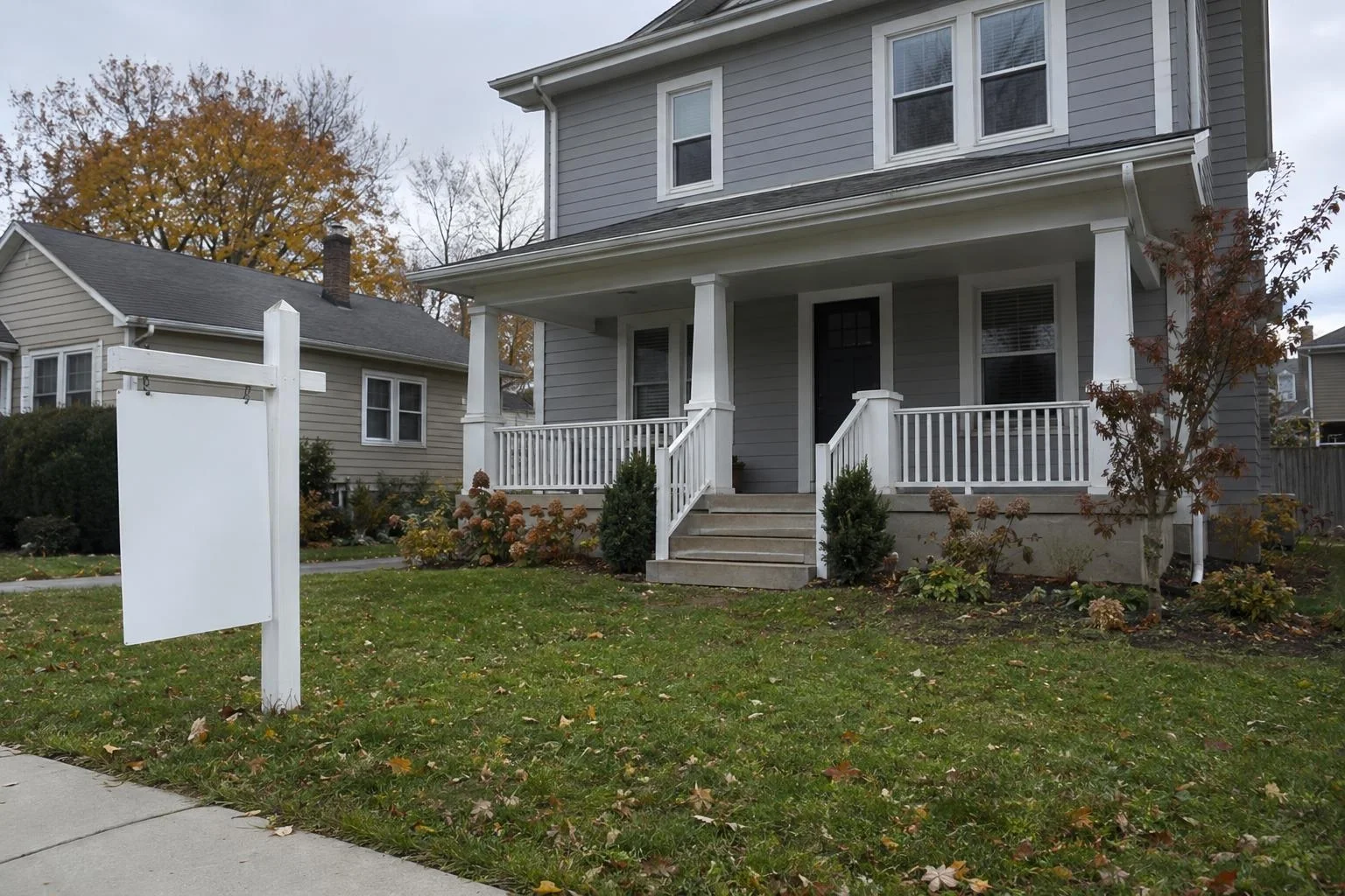 A gray two-story house with a front porch and a blank white real estate sign on the lawn in autumn