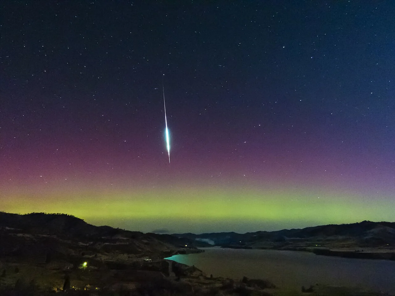 A bright green-white fireball streaks down a starry sky above dark hills and a lake, with a band of green and pink aurora along the horizon