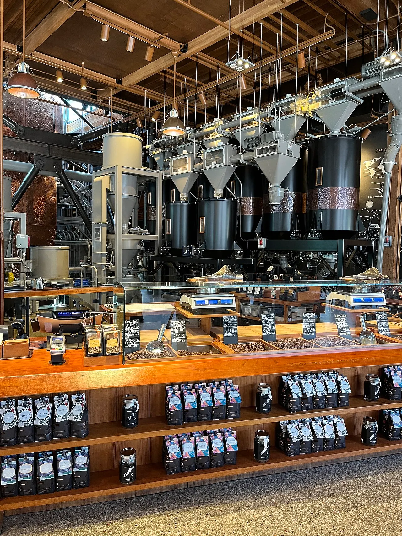 Bins of coffee beans with chalkboard price signs and bagged coffee on shelves at a Starbucks roastery counter, with roasting silos behind