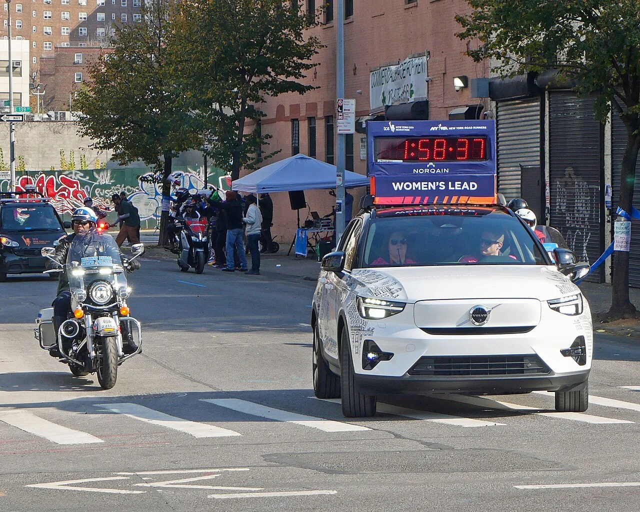A police motorcycle and the women's lead timing car on a Bronx street, with spectators under a tent