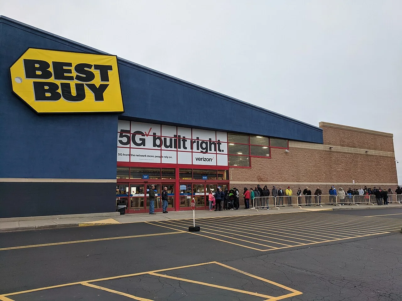 Shoppers wait in a line along a barricade outside a Best Buy store before it opens on an overcast morning