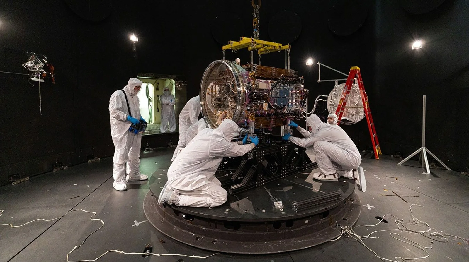 Engineers in white cleanroom suits work around a spacecraft on a round platform inside a large dark test chamber
