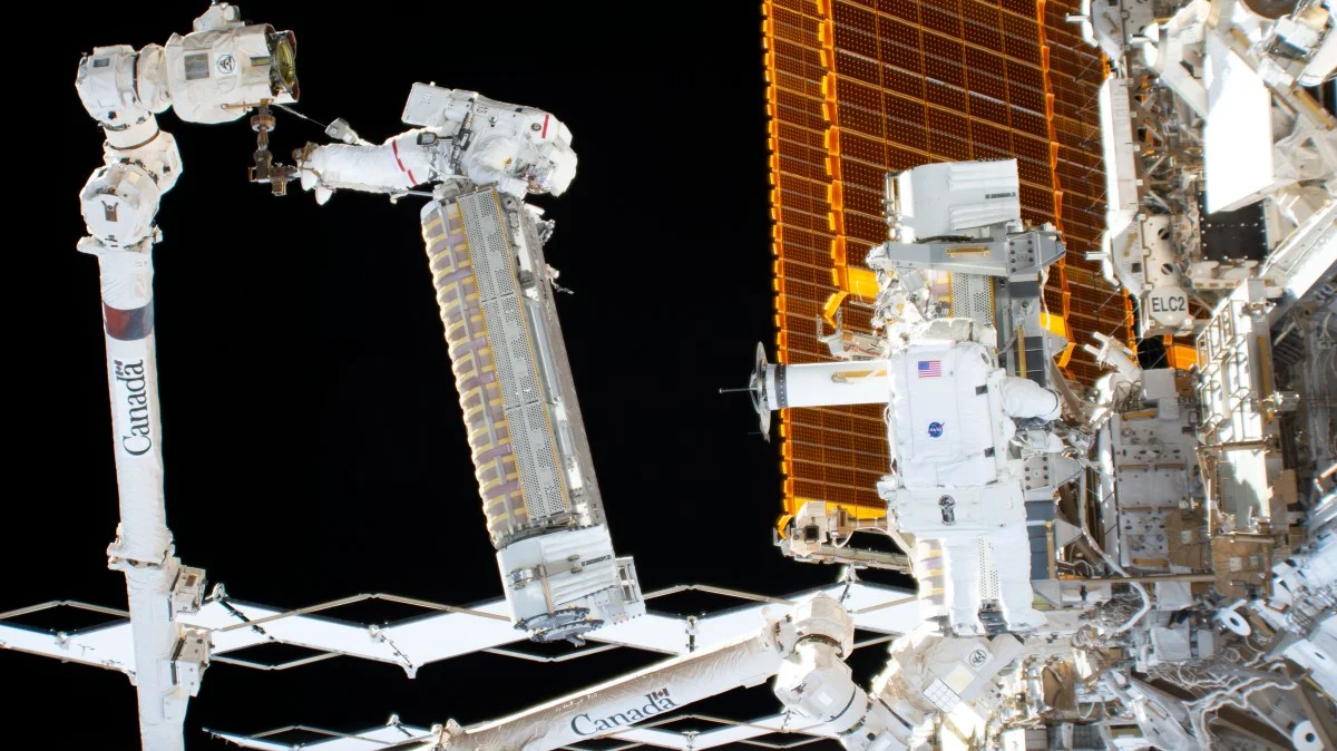 Two astronauts and Canadarm2 handle a folded roll-out solar array beside older orange solar wings