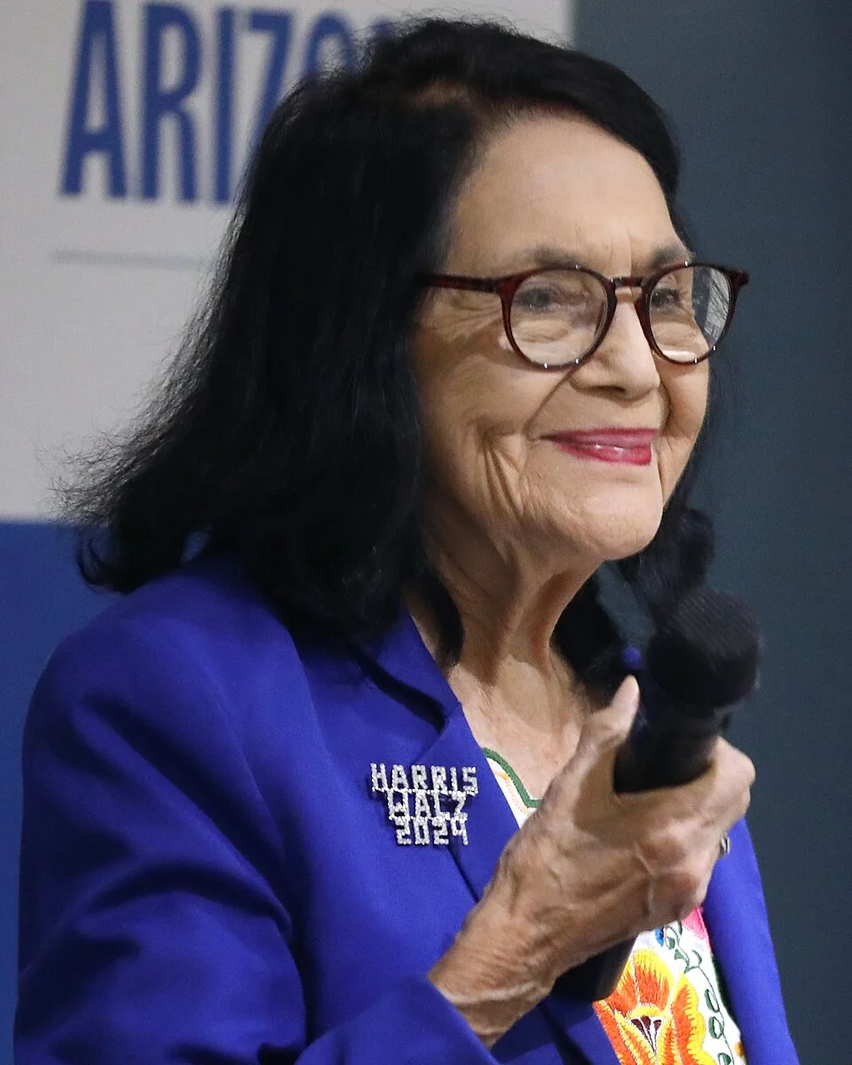 Dolores Huerta, in glasses and a blue jacket, smiles while holding a microphone at a press conference in Phoenix