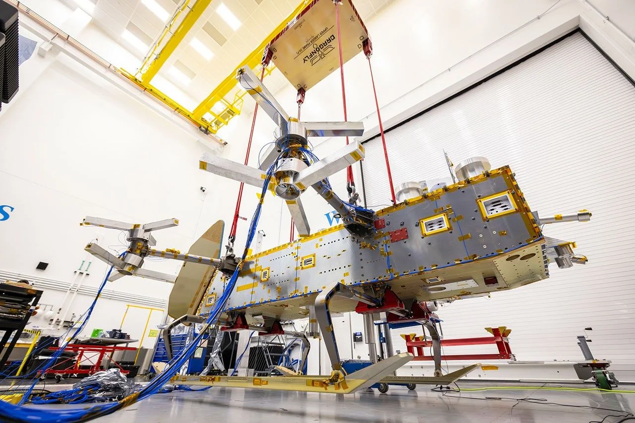 The Dragonfly rotorcraft, a long silver body with rotor arms and landing skids, hangs from red cords a few inches above the floor of a clean room