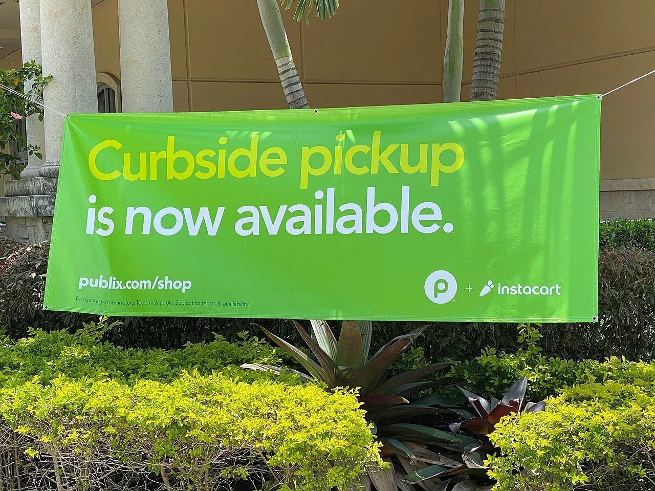 A green Publix and Instacart banner reading Curbside pickup is now available, hung above shrubs outside a store
