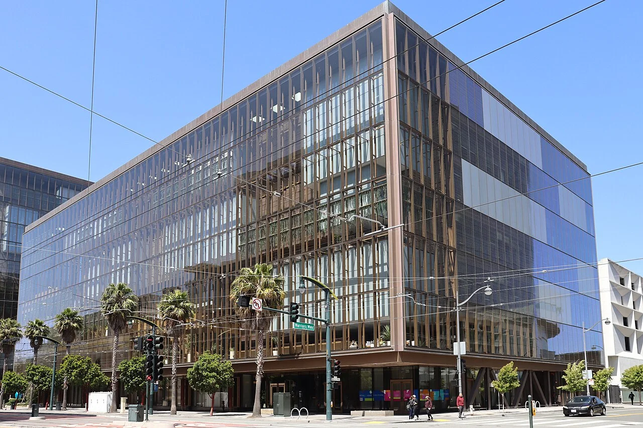 A glass-walled office building on a San Francisco street corner with palm trees and streetlights