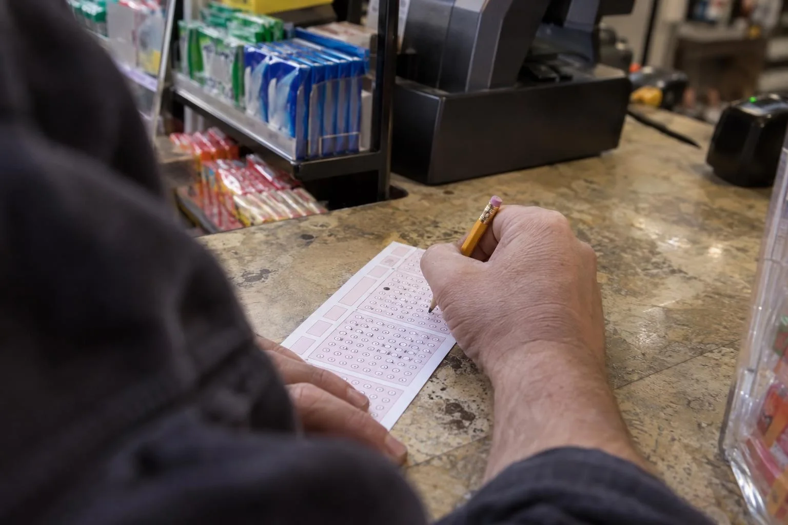 A hand fills in bubbles on a lottery play slip with a pencil at a store counter