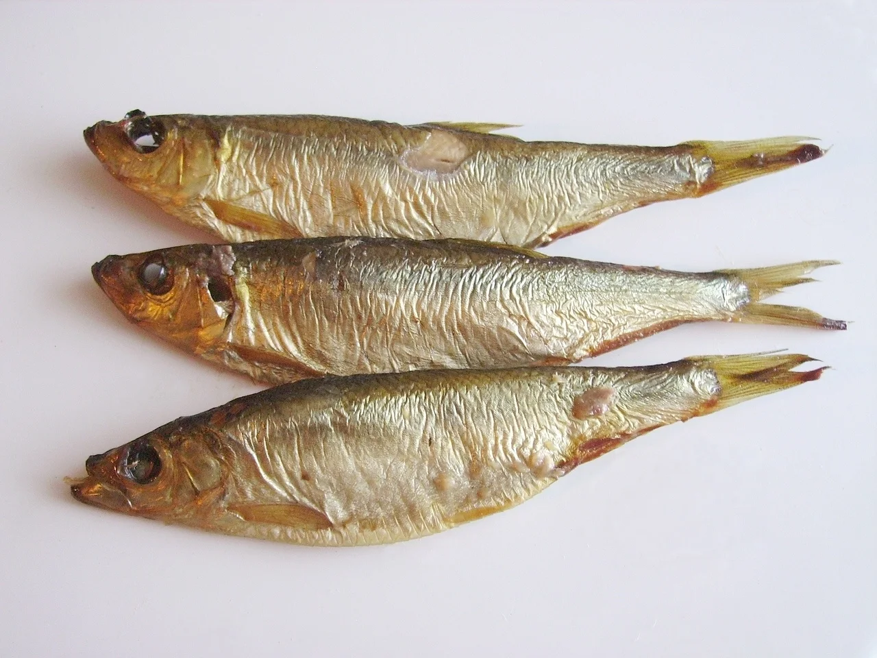 Three whole smoked European sprats on a white surface in an archival photograph.