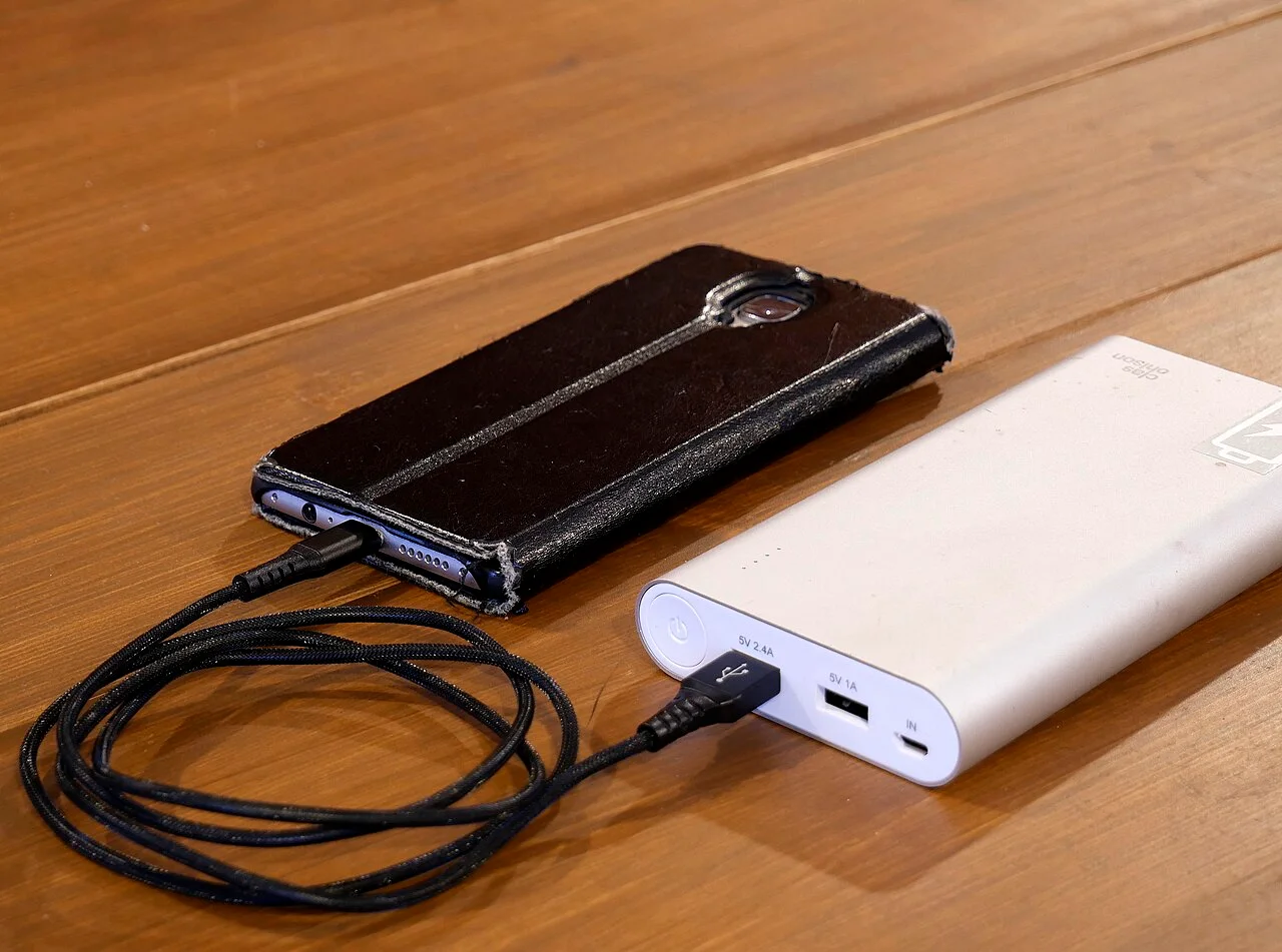 A silver power bank on a wooden table connected by a black cable to a smartphone in a black case