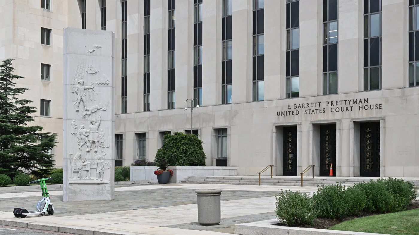 Front of a stone federal courthouse with the words E. Barrett Prettyman United States Court House over its doors and a carved stone column on the plaza