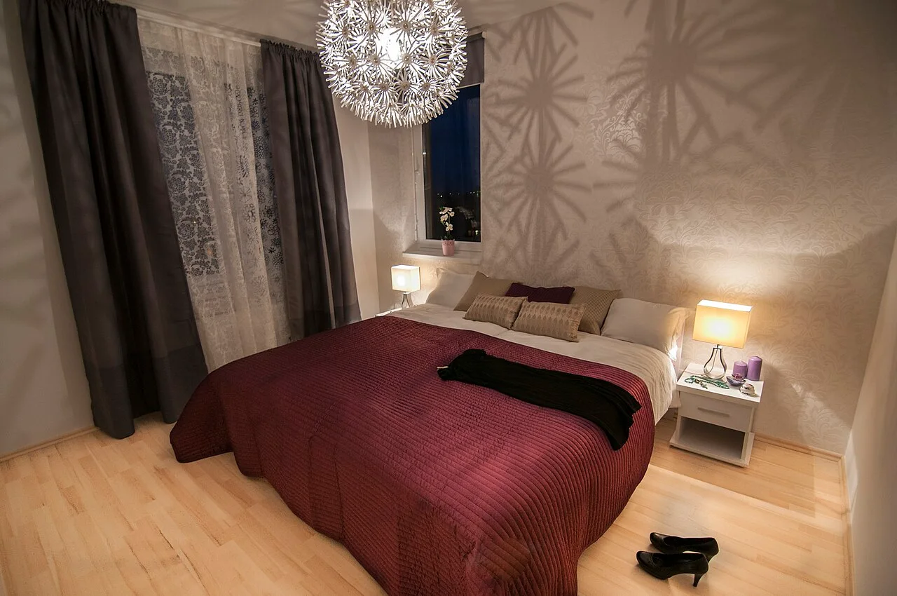 A bedroom with a burgundy bedspread and bedside lamps casting shadows on the wall.