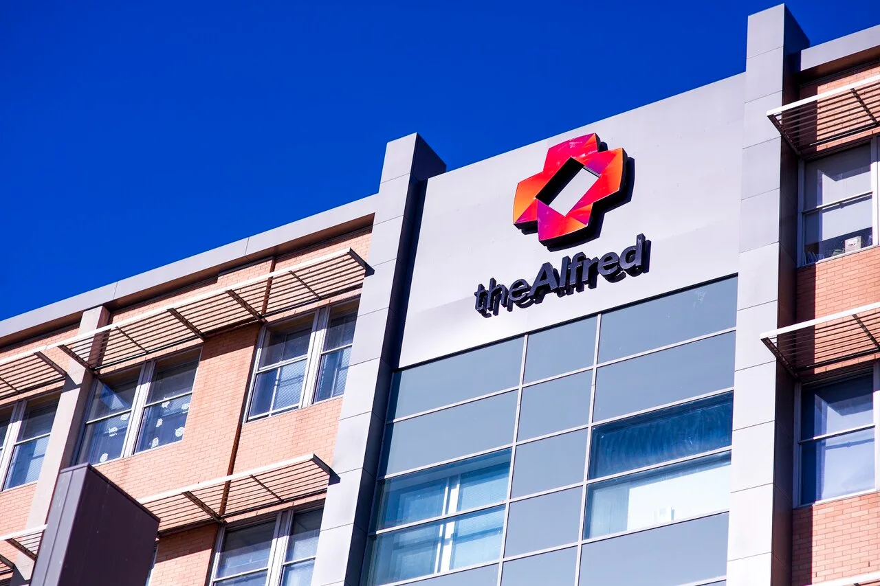 Upper floors of a brick and glass hospital building with a red and orange logo and the name theAlfred against a blue sky