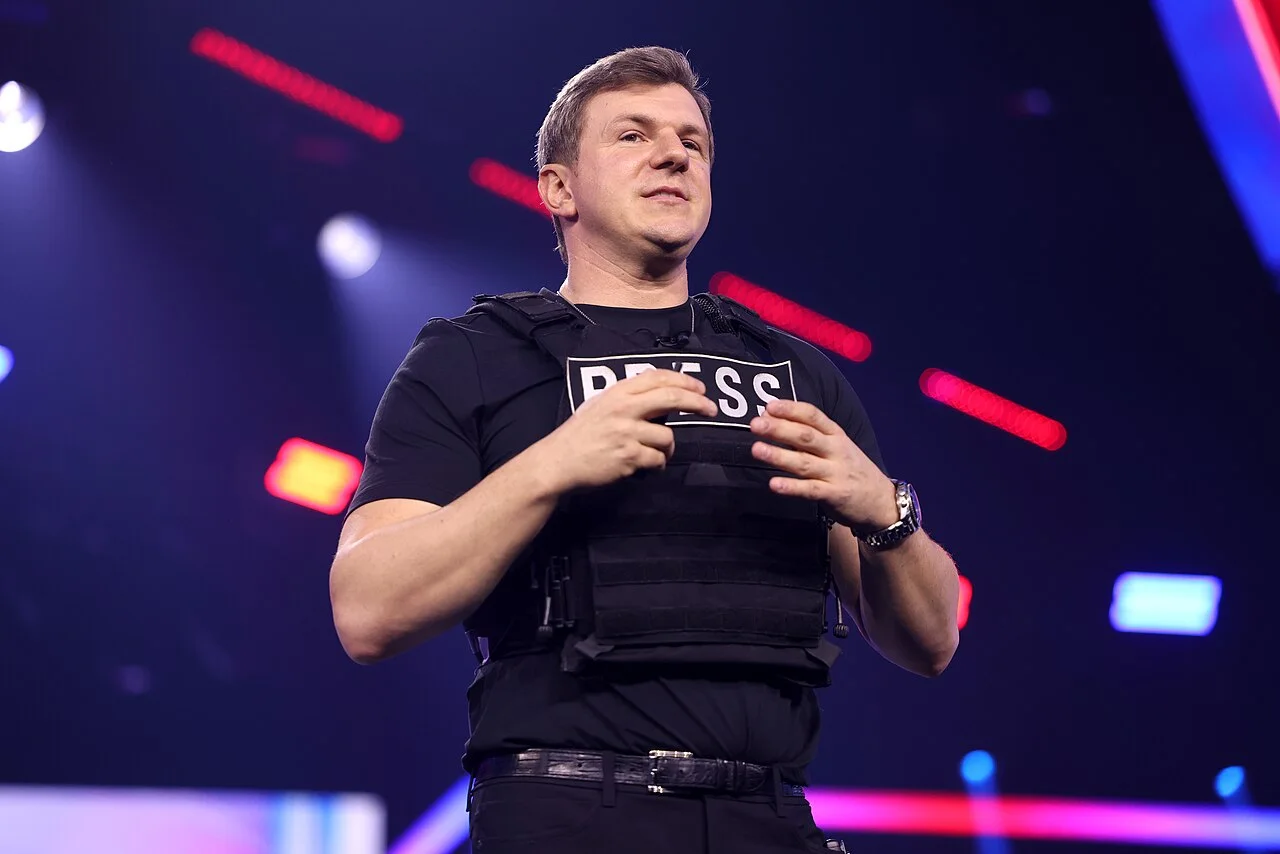 James O'Keefe in a black vest marked PRESS speaking on a lit stage