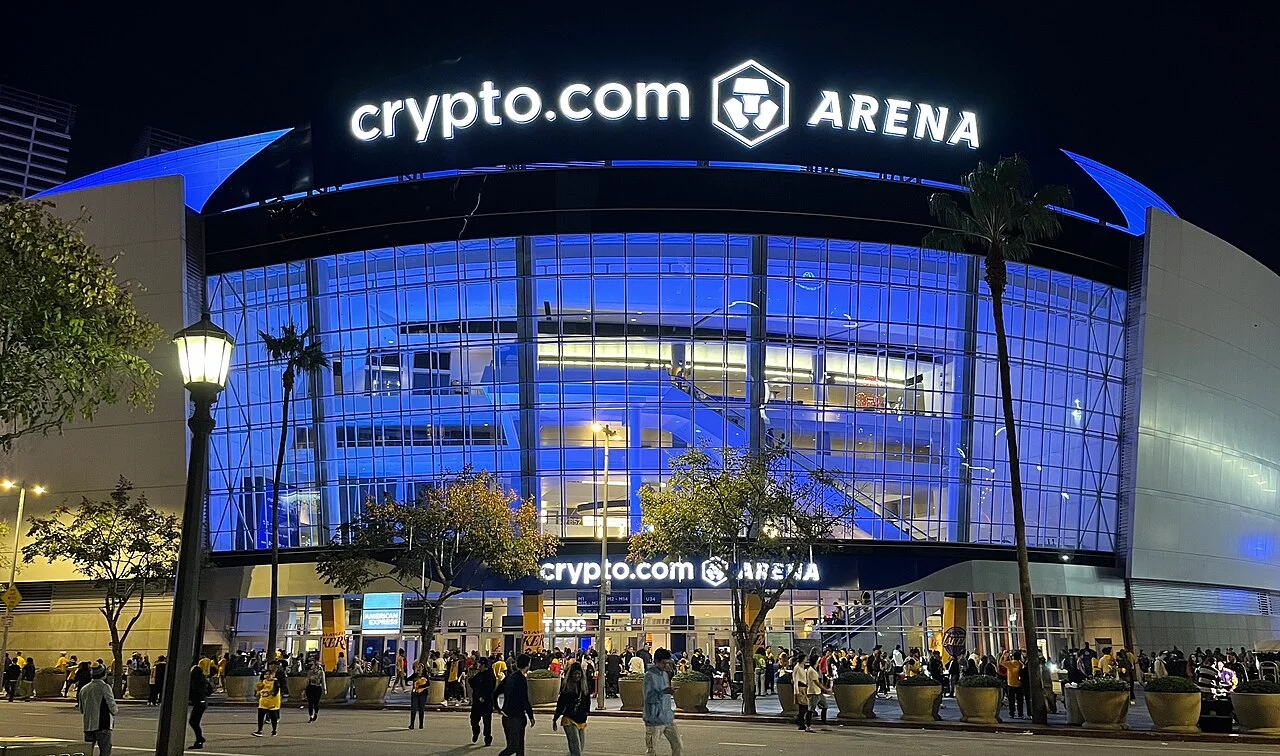 The glass front of Crypto.com Arena lit blue at night, with crowds on the plaza and the arena name in lights above