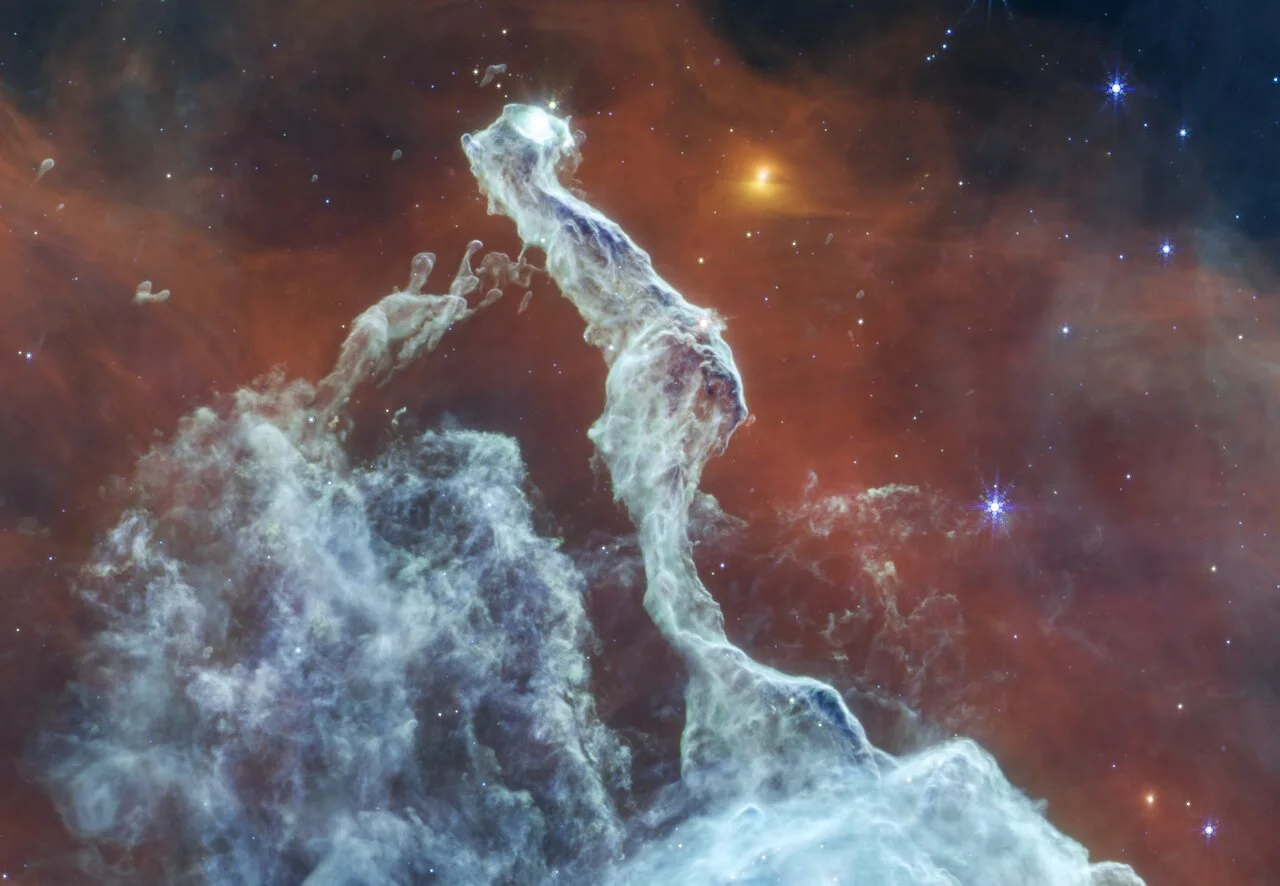 Webb mid-infrared image showing a pale, narrow dust pillar and fine strands against a reddish background, with a yellow source to the right.