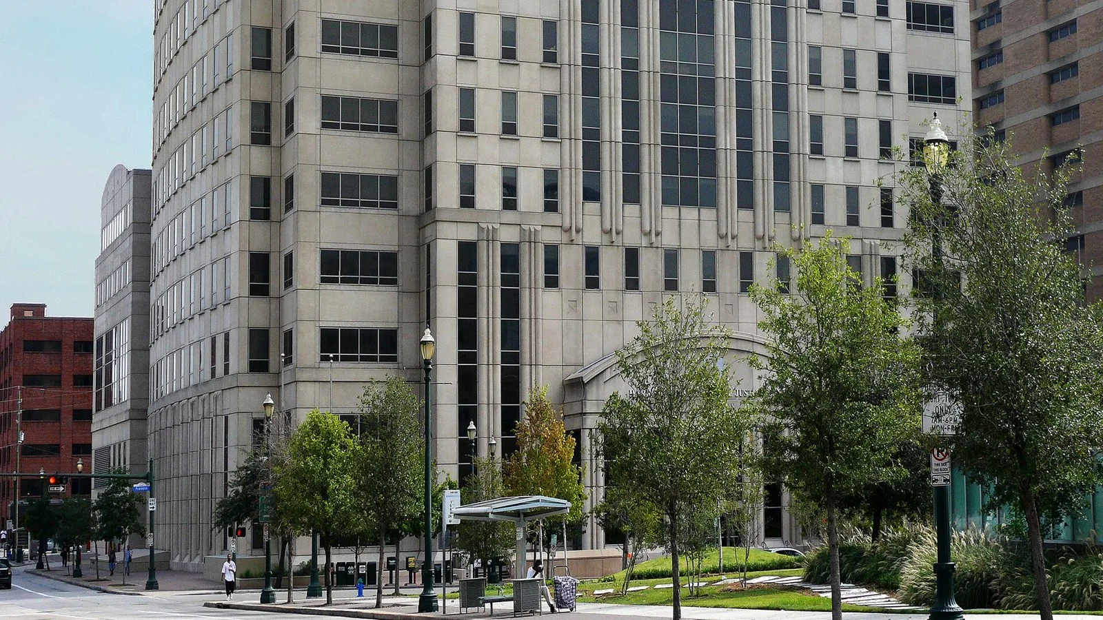 A tall stone-clad courthouse tower with an arched entrance behind young trees and a bus shelter on a downtown Houston street