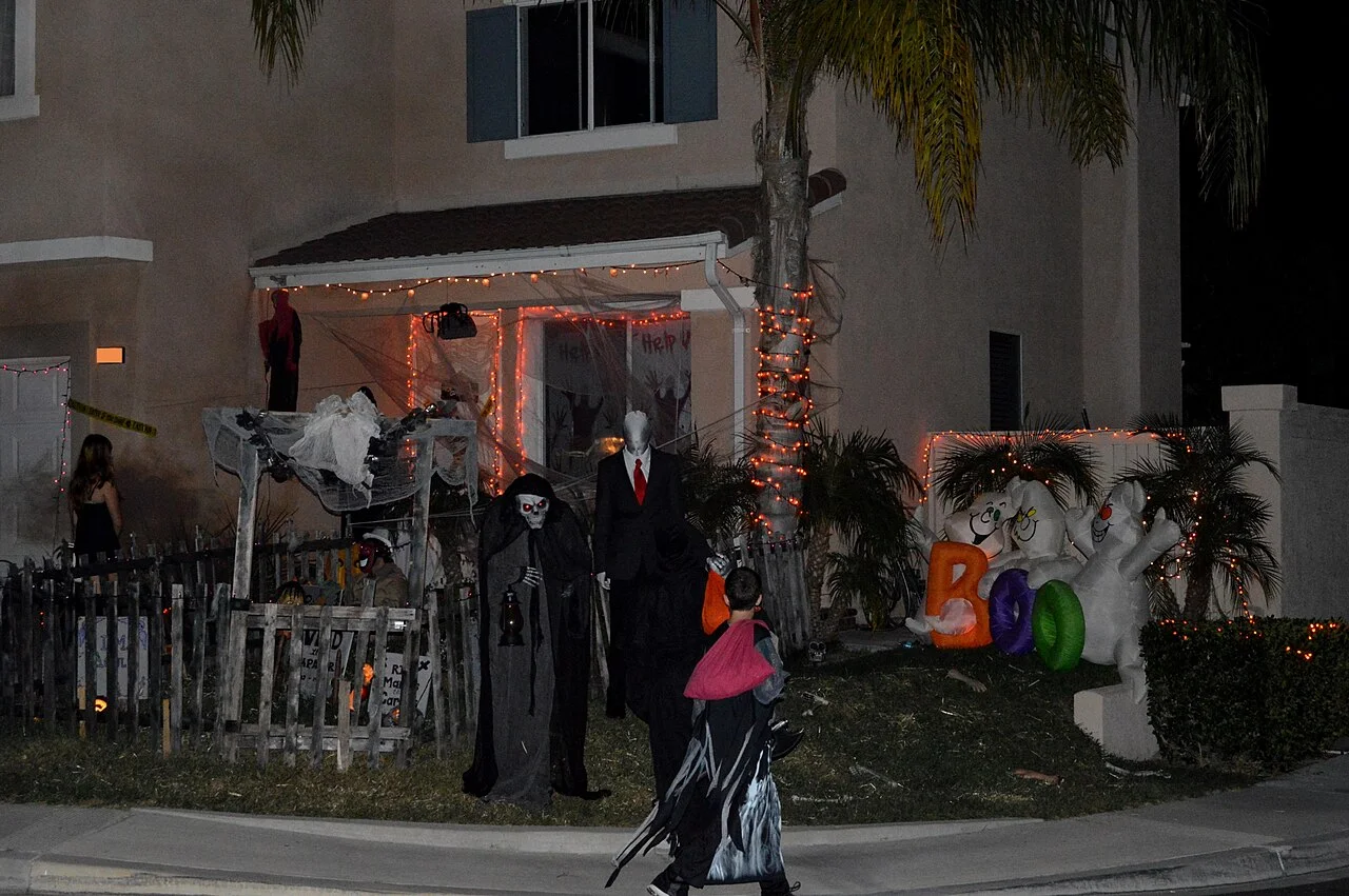 A child in a dark costume walks past a house decorated with Halloween lights, skeleton figures and inflatable ghosts at night
