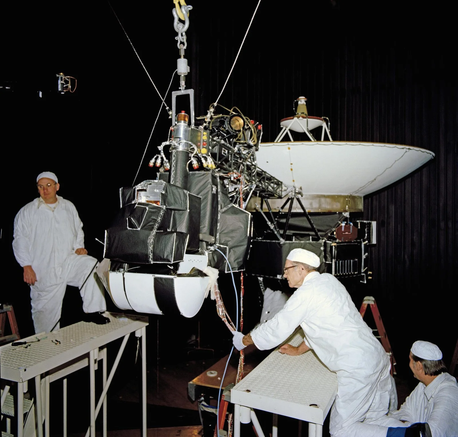 Technicians in white clean-room suits work around a Voyager spacecraft with its large white dish antenna inside a dark test chamber