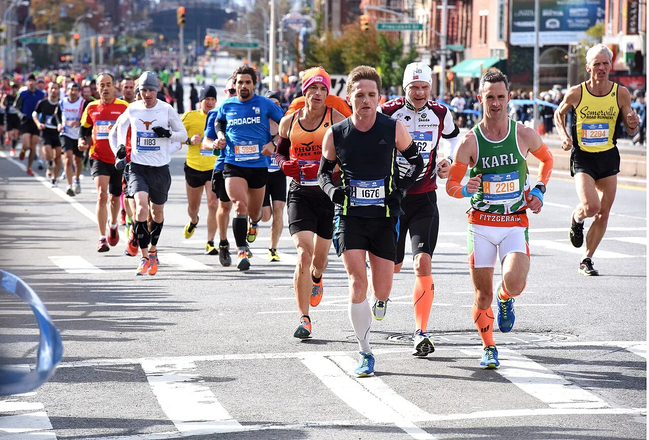 Marathon runners with race bibs running down a wide Brooklyn avenue, with spectators behind barriers on the sidewalk