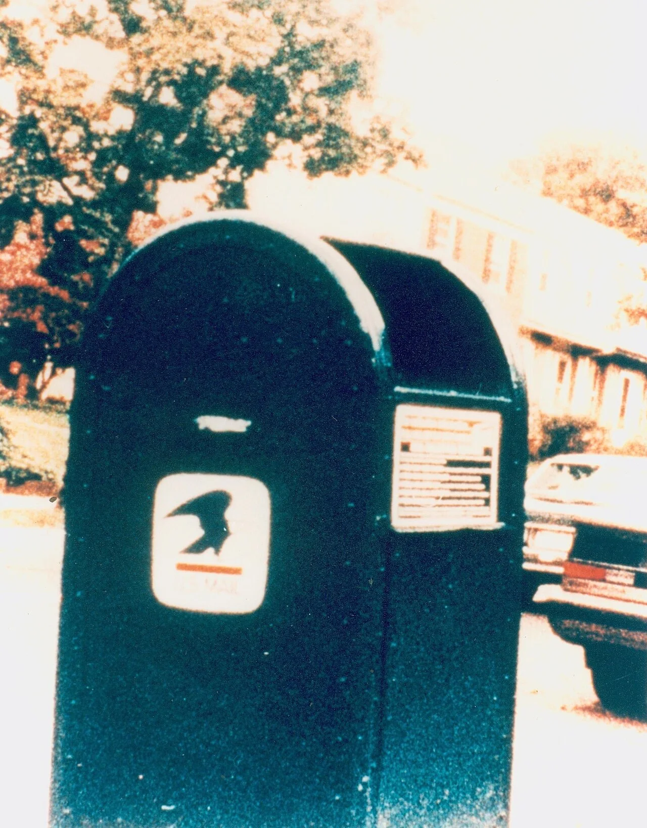 A blue U.S. mailbox on a residential street with a small horizontal mark above the postal eagle emblem