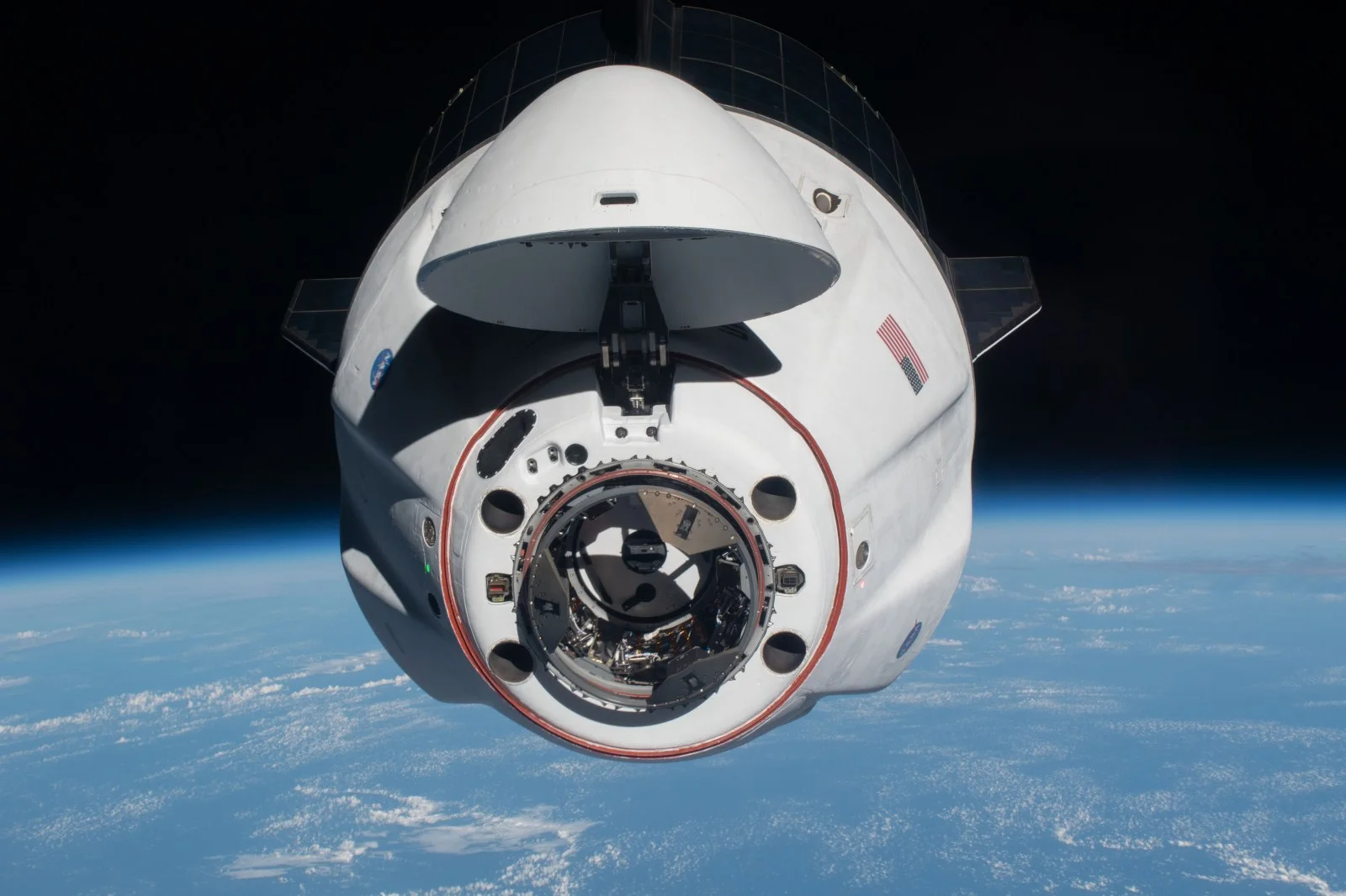 A white SpaceX Crew Dragon capsule with its nose cone open, approaching above the Earth