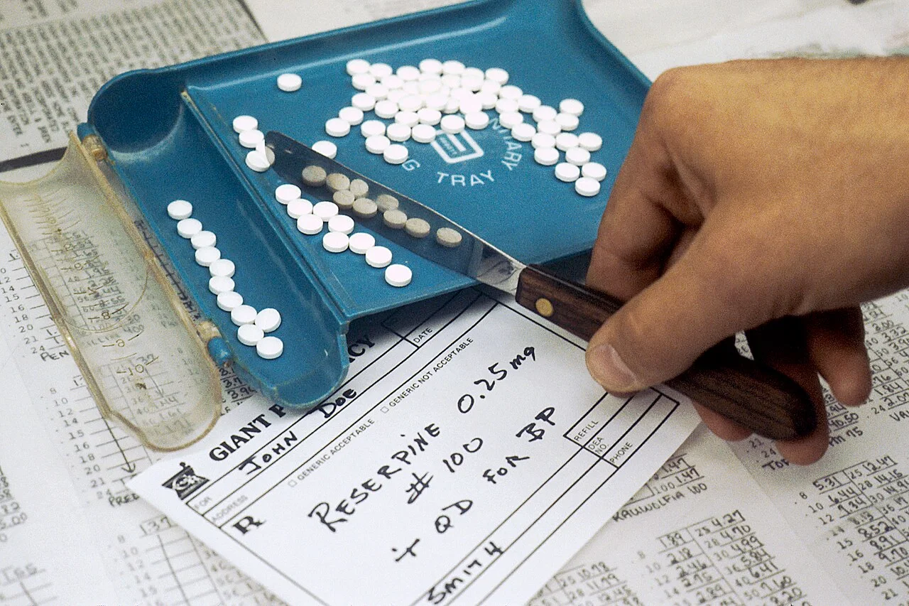 A hand uses a spatula to count white prescription tablets on a blue pharmacy tray.