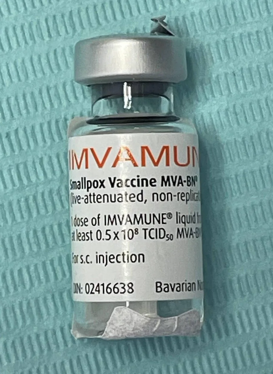 A small glass vaccine vial with a silver cap and a label reading Imvamune Smallpox Vaccine MVA-BN, on a blue cloth
