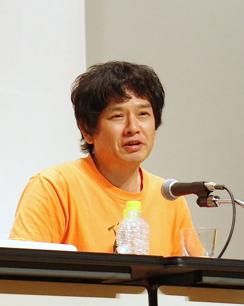 Yoshitomo Nara in an orange shirt speaking into a microphone at a press conference in Yokohama in July 2012.
