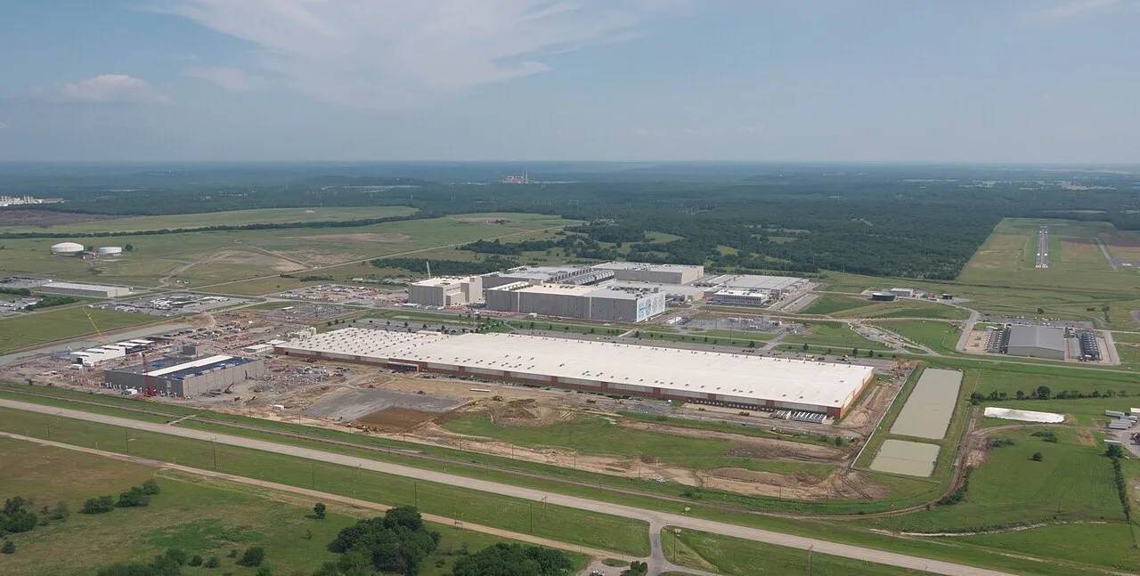 Aerial view of a data center campus with large white-roofed buildings, construction areas and roads among green fields and woods