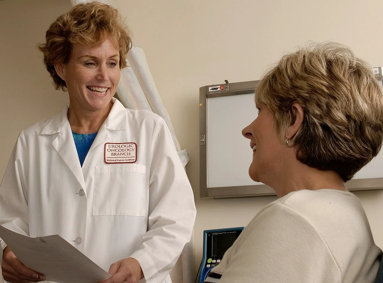 A doctor holding a clipboard talks with a patient in a medical examination room.