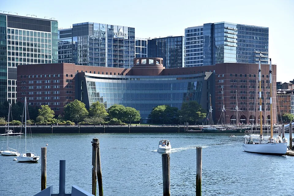 The brick John Joseph Moakley federal courthouse in Boston with its curved glass facade, seen across the harbor with sailboats in front