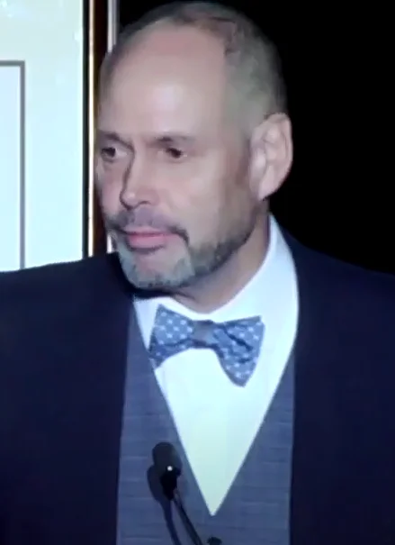 Ernie Johnson in a dark jacket, sweater vest and blue bow tie speaking at a microphone