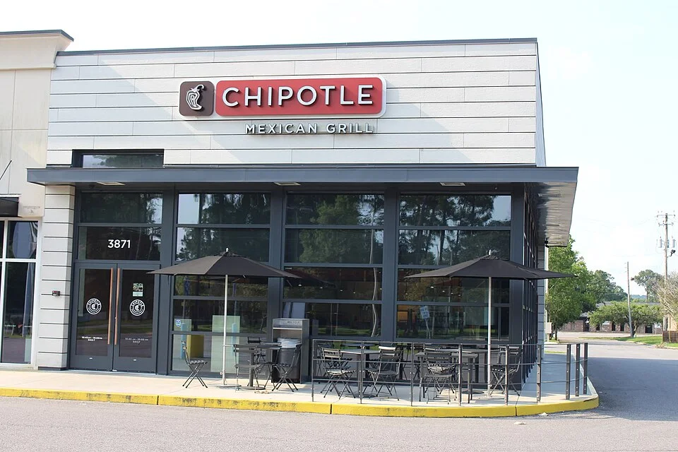 A Chipotle Mexican Grill storefront with a red sign, glass front and patio tables under umbrellas