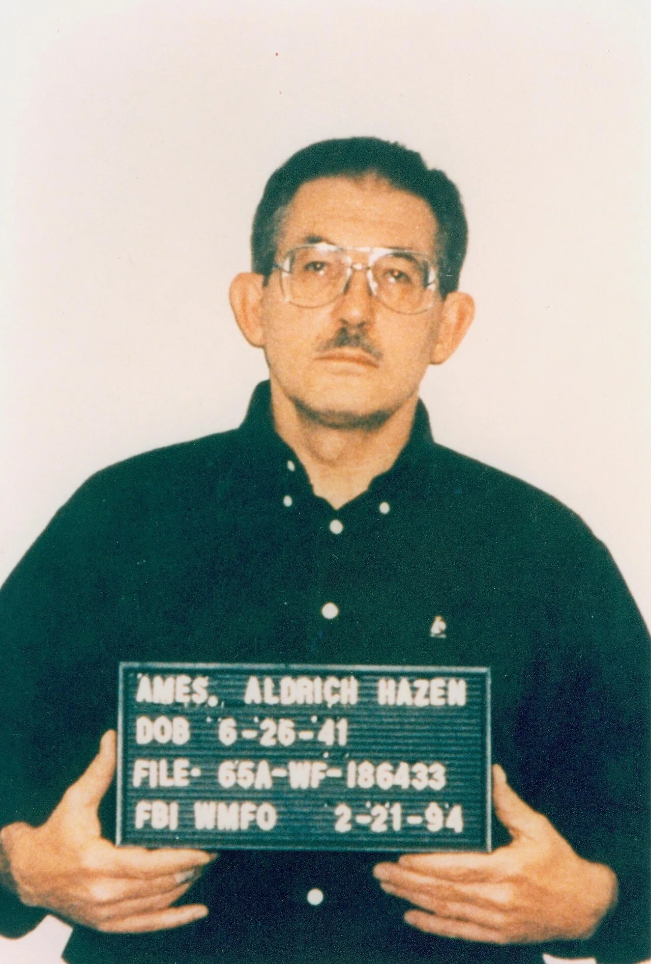 Aldrich Ames in glasses and a dark shirt holding an FBI booking placard dated 2-21-94