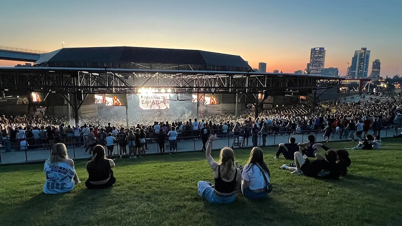Fans sit on a grass lawn facing a covered outdoor amphitheater stage at dusk, with the Milwaukee skyline behind