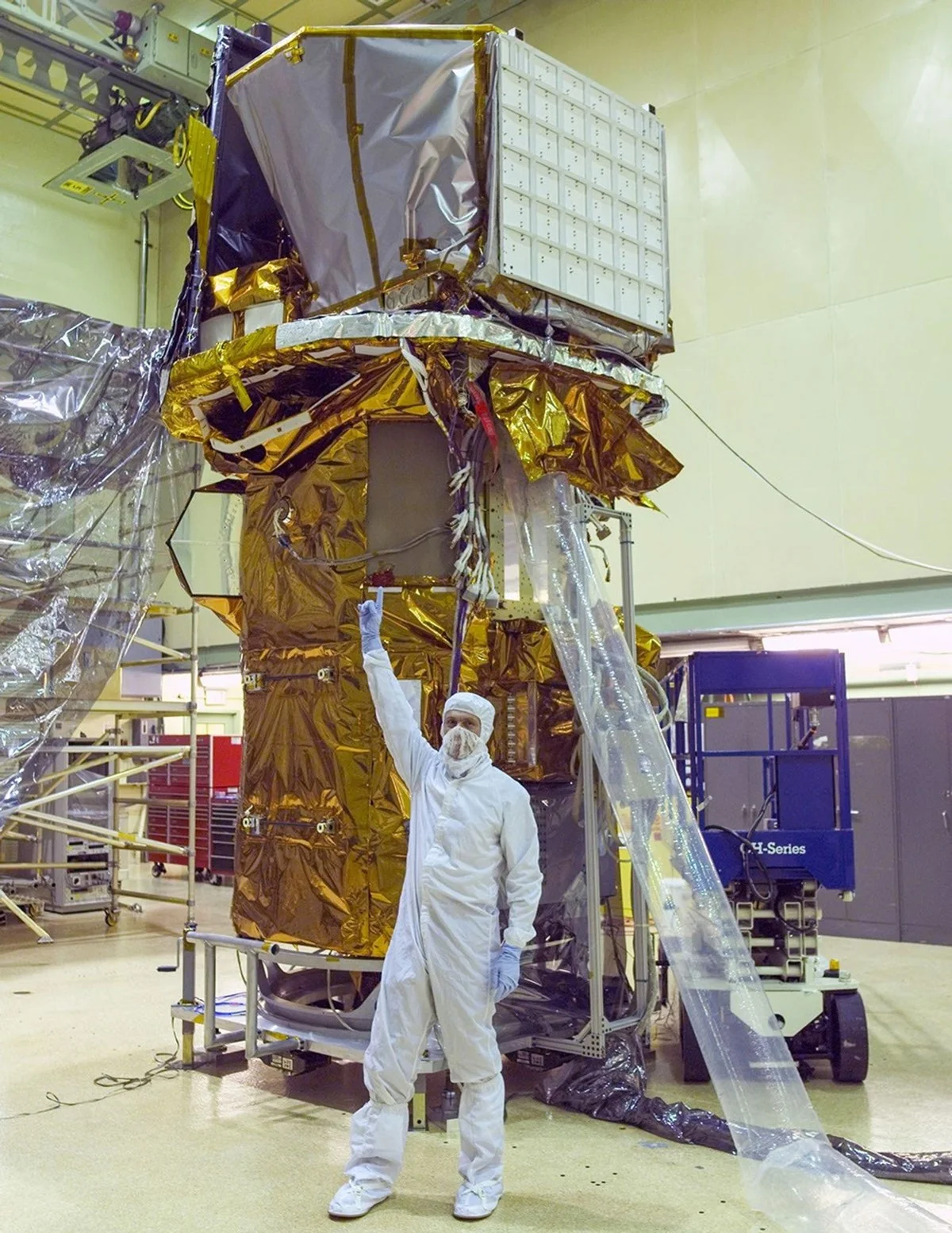 A man in a white cleanroom suit points up at a tall spacecraft wrapped partly in gold foil