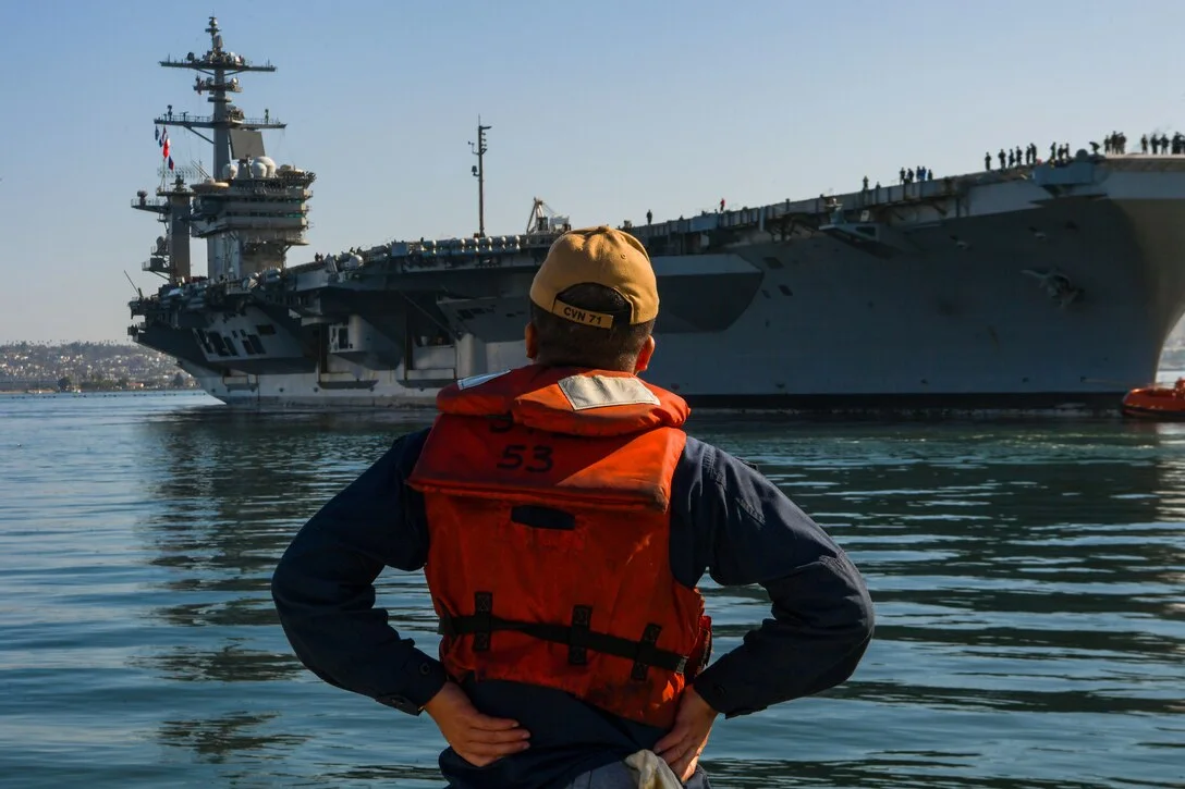 USS Abraham Lincoln approaches its mooring in San Diego in July 2025, seen behind a Navy sailor on a boat