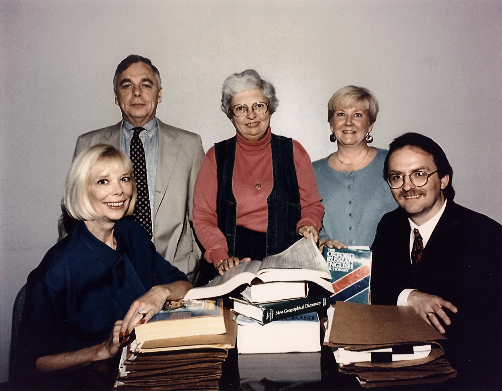 Five CIA officers, two seated and three standing, posed around a table stacked with files and reference books